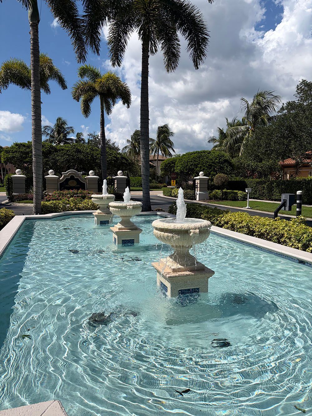 Blue fountain pool with three tiered water jets, surrounded by palm trees and manicured gardens under cloudy skies