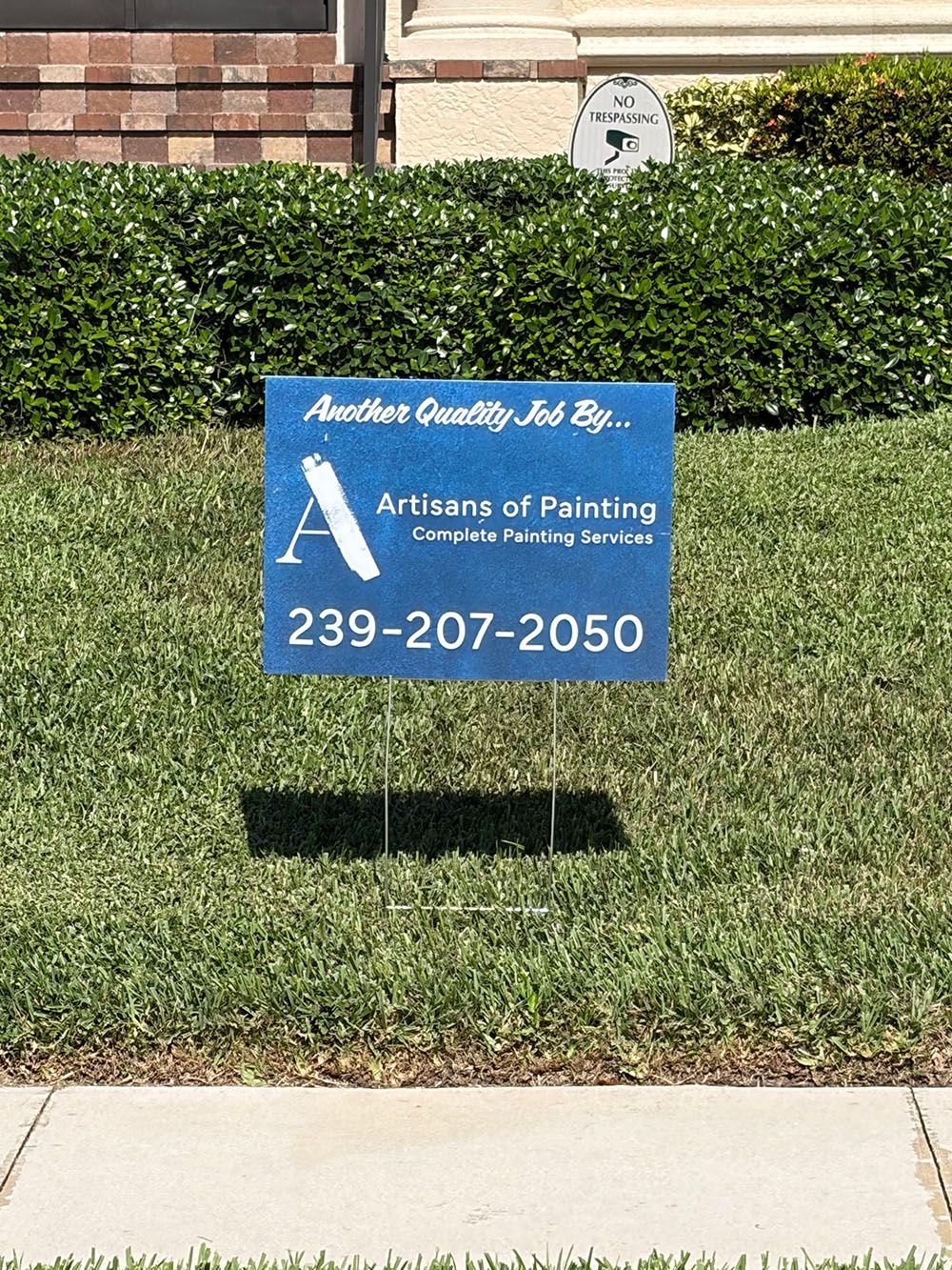 Blue lawn sign for “Attitudes of Painting” with phone number 239-207-2050 in front of a hedge and building