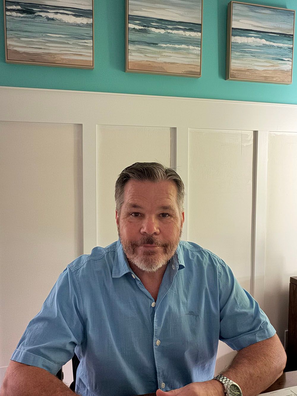 Man in a blue shirt seated at a desk in a bright room with teal wall art and white cabinet doors.