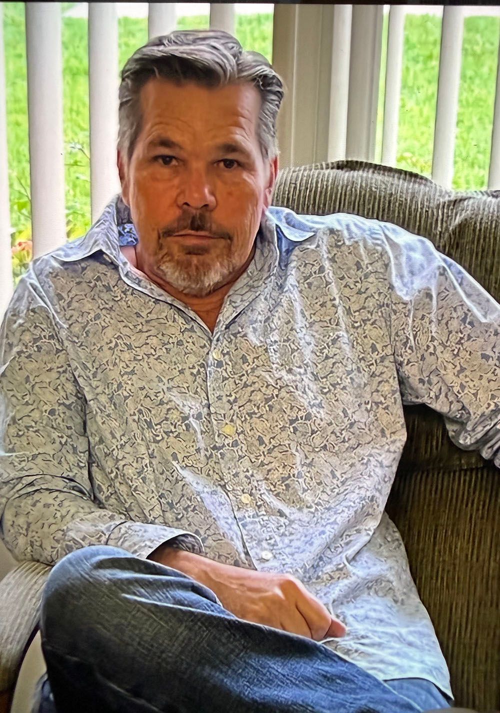 Man sitting on a couch by a window, wearing a light patterned shirt and looking at the camera.