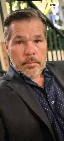 Close-up of a man in a dark blazer and patterned shirt, seated outdoors with greenery behind him