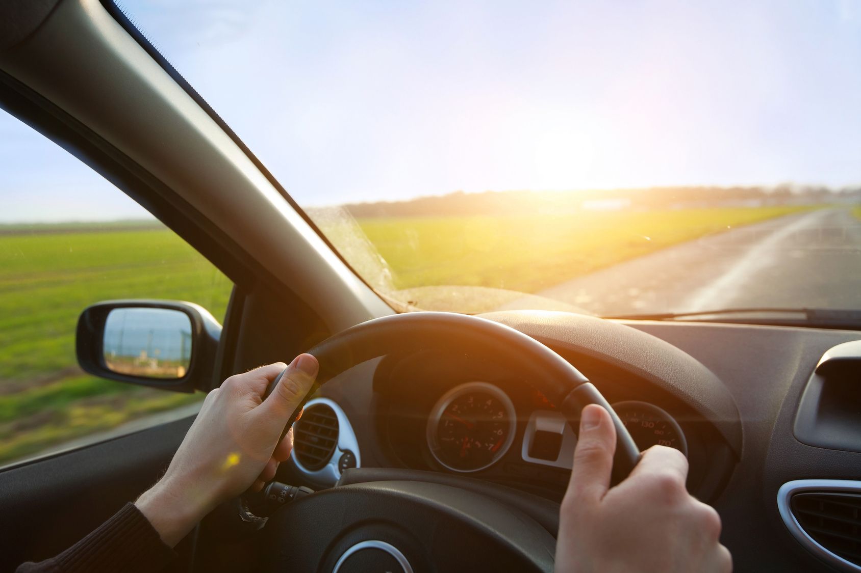 Hands on steering wheel driving toward a sunlit road beside green fields at sunset