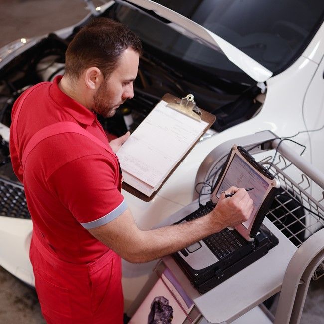 Mechanic in red uniform inspecting a car engine with a diagnostic laptop and clipboard