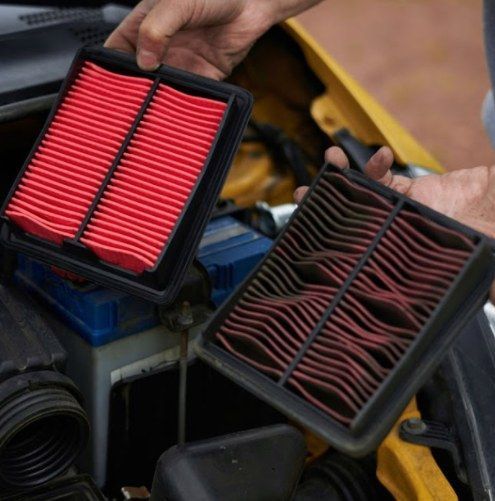 Hands holding two car air filters, one red pleated and one dark dirty filter, above engine parts.