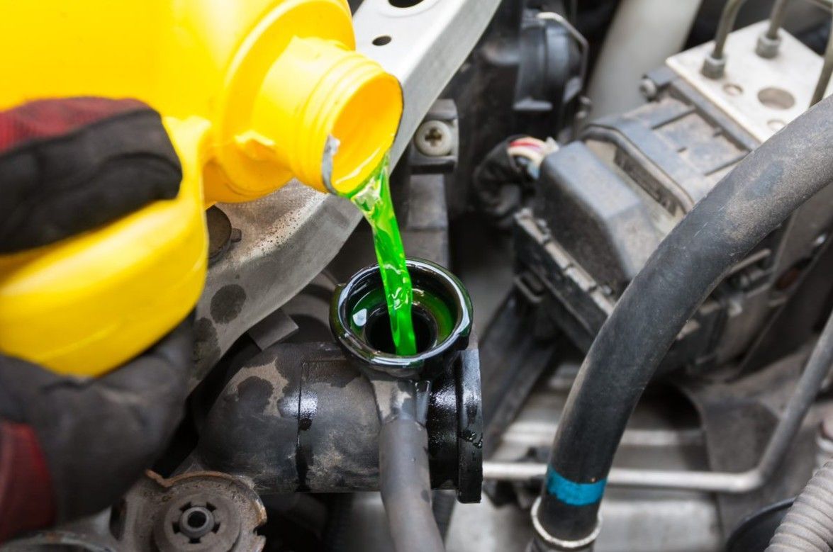 Gloved hand pouring green coolant from a yellow bottle into a car engine reservoir