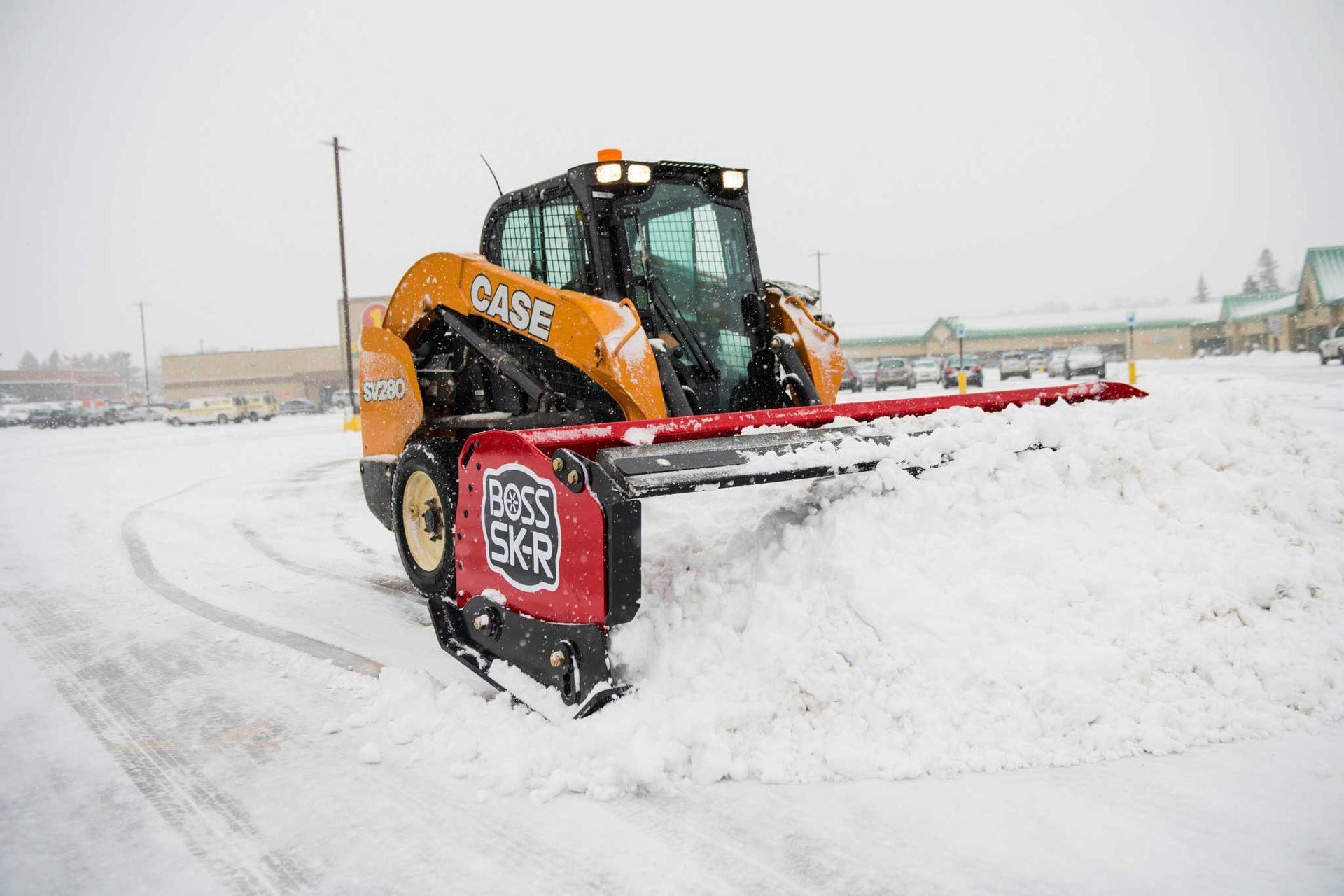 A Case skid steer with a snow blade clears a snowy parking lot. It’s a winter scene with a red and orange machine.