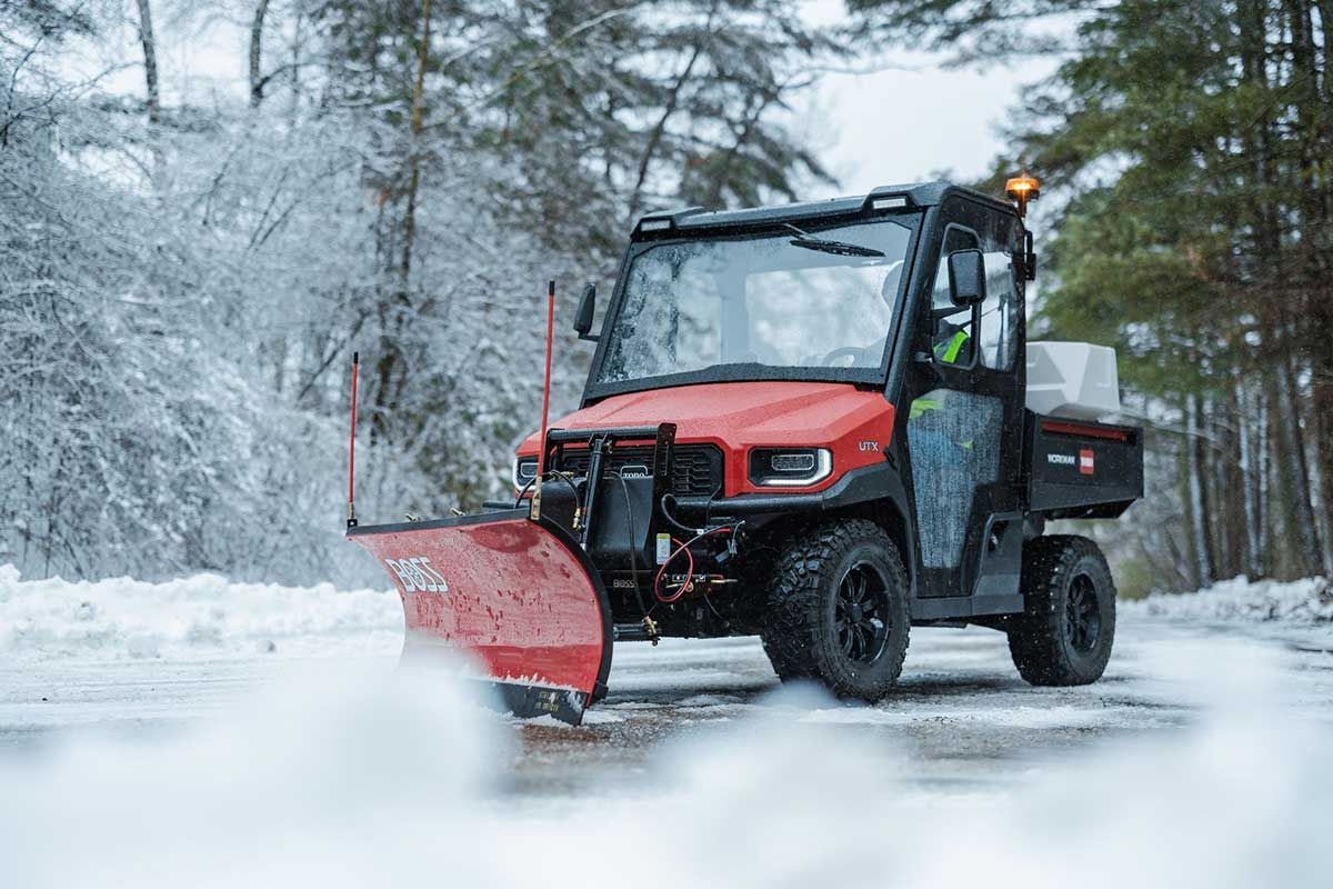 A Toro utility vehicle plows snow from a paved road. The red snowplow blade pushes white snow aside, with trees in the background.