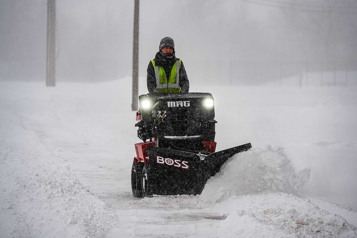 Person operating a red Boss snowblower, clearing a snowy sidewalk in a blizzard. The operator wears a reflective vest and a hat.
