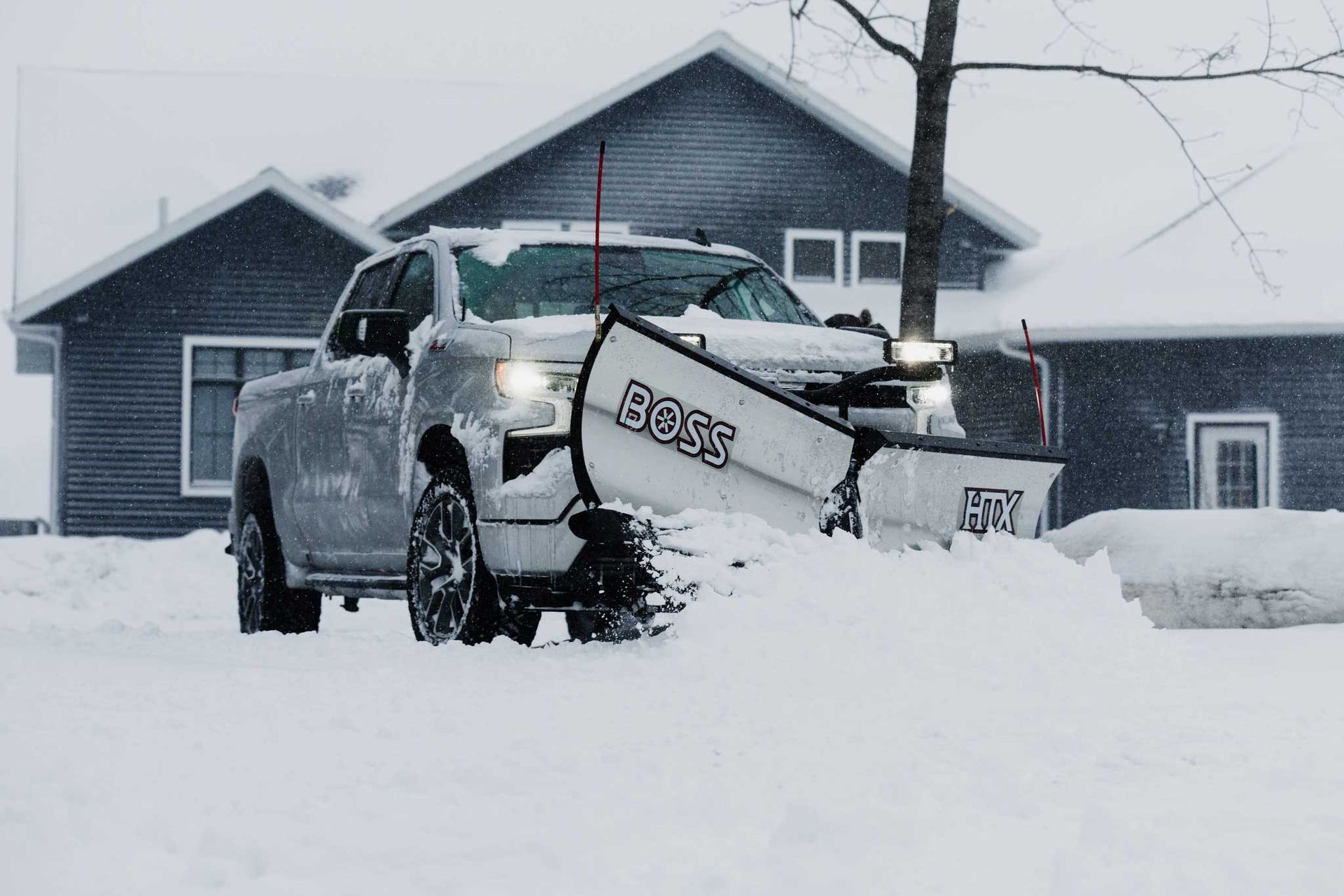 A silver pickup truck with a snowplow clearing snow in front of a house during a snowstorm.