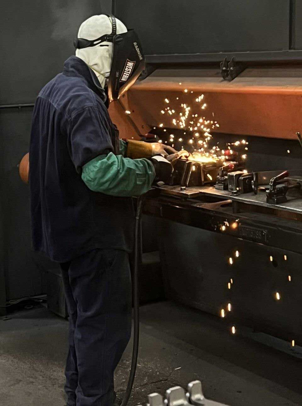 Welder wearing a protective mask and gear, sparks flying from the metal he is working on in a workshop.
