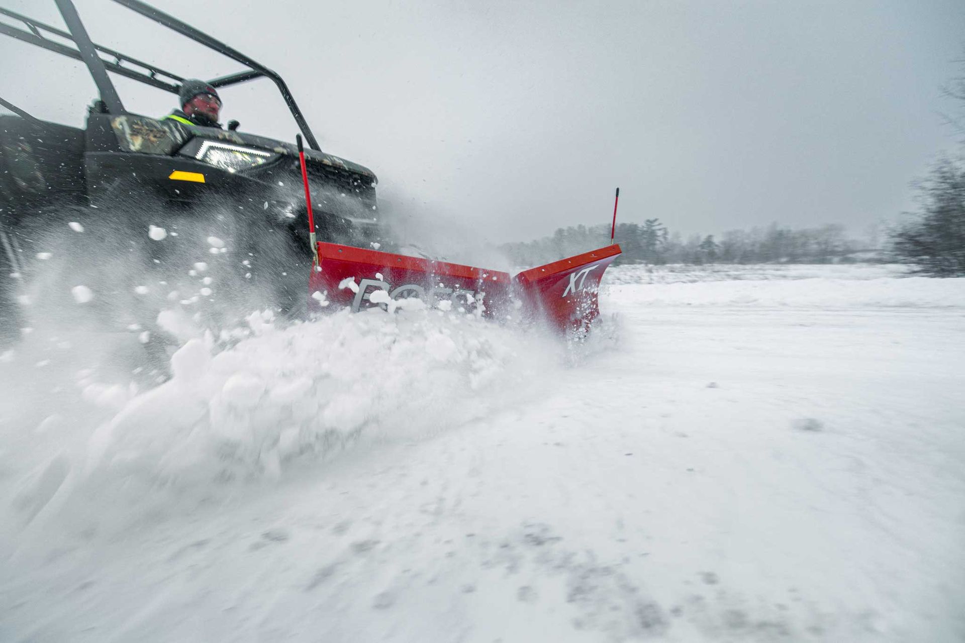 An ATV plows through snow, pushing it to the side with a red blade. The driver is visible inside the vehicle.