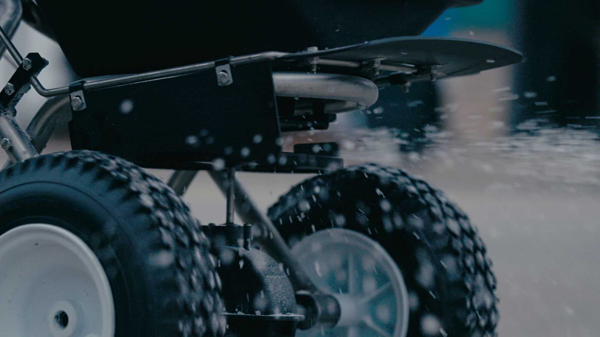 Close-up of a spreader spraying white material onto a surface. The spreader has two large wheels.