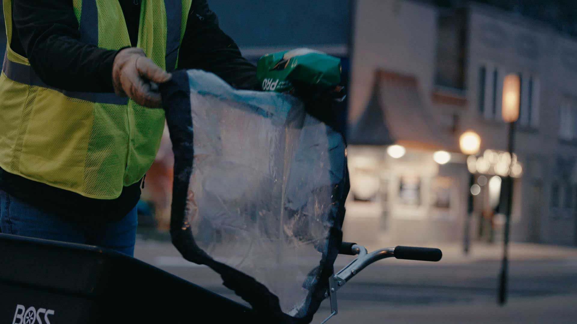 Person in a reflective vest holding a clear plastic bag, preparing to load it onto a cargo bike at dusk.