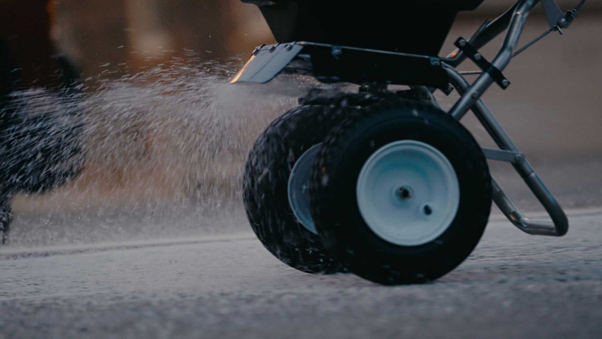 A person spreading salt on a road with a push spreader; white salt sprays out.