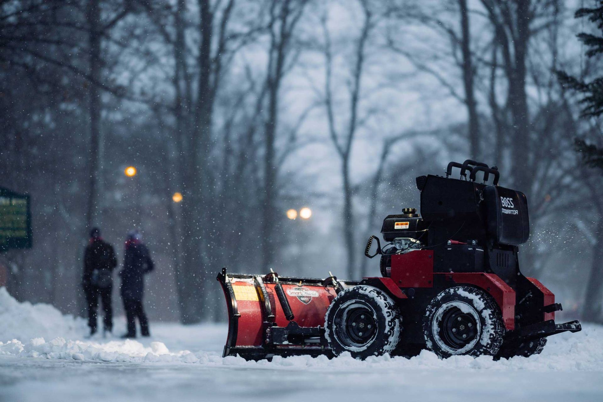 A red snowplow clearing a snowy path, with two figures watching in the background; trees and streetlights are visible.