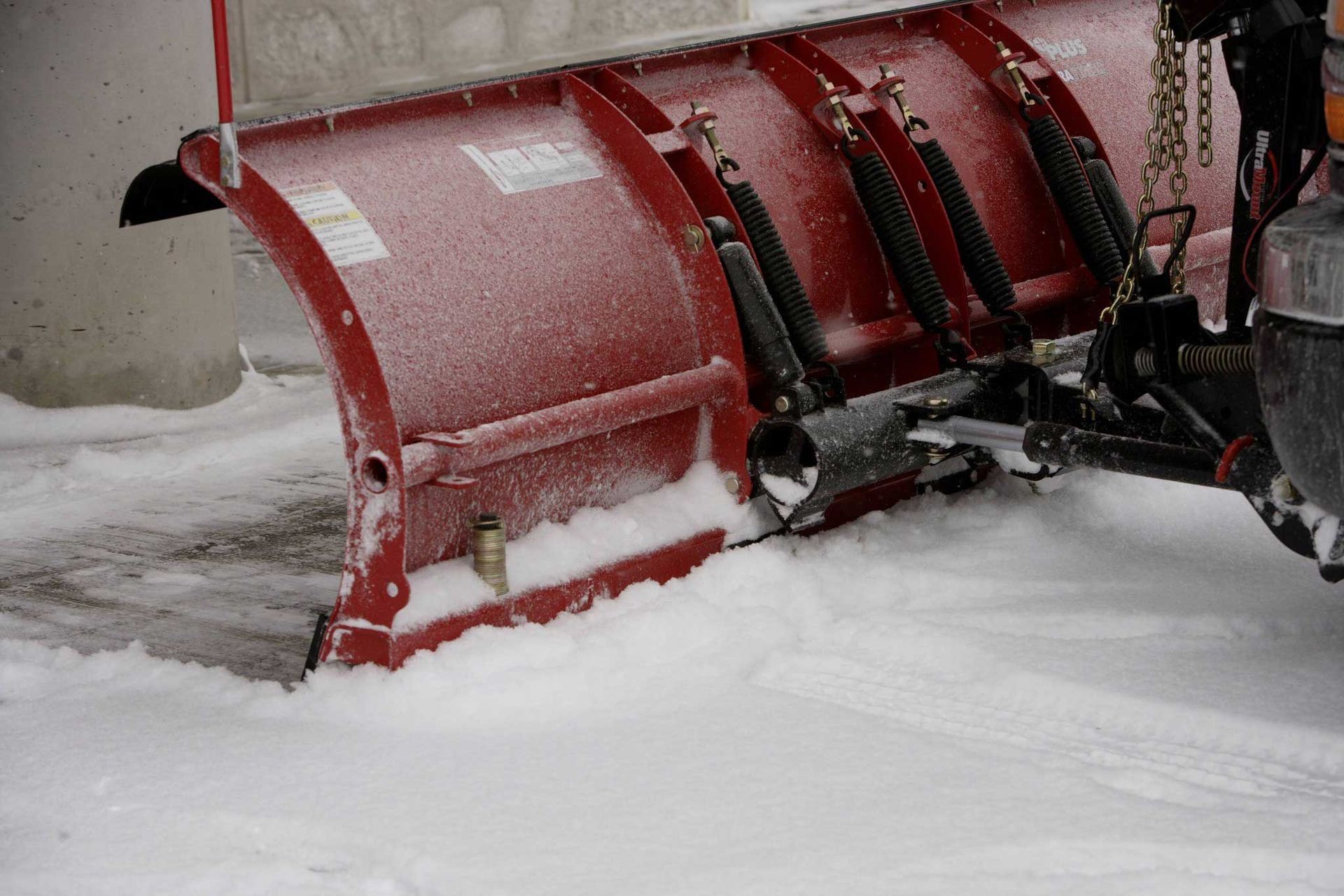 Red snowplow clearing snow from a driveway. The plow is attached to a vehicle, with snow piled up in front of it.