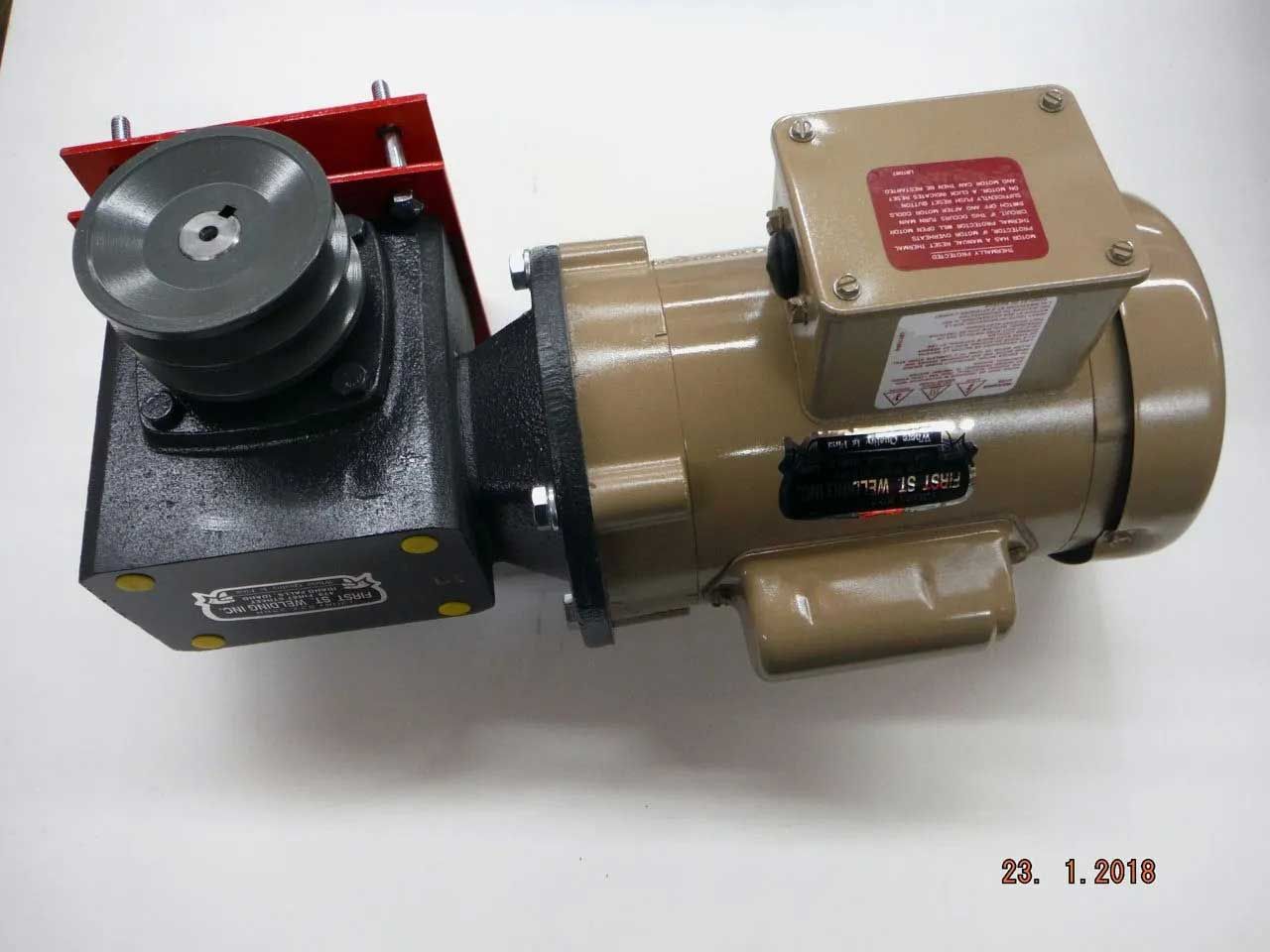 An electric motor with a gearbox and pulley system, mounted on a metal plate. The motor and gearbox are tan and black, respectively.