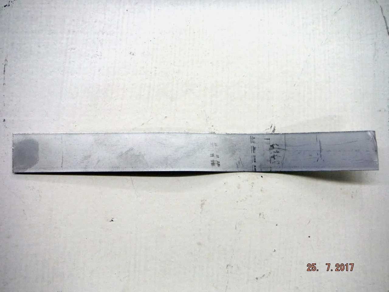 A metal strip with handwritten markings, likely for measurement or testing, lies on a white surface. Date stamp present at the bottom.