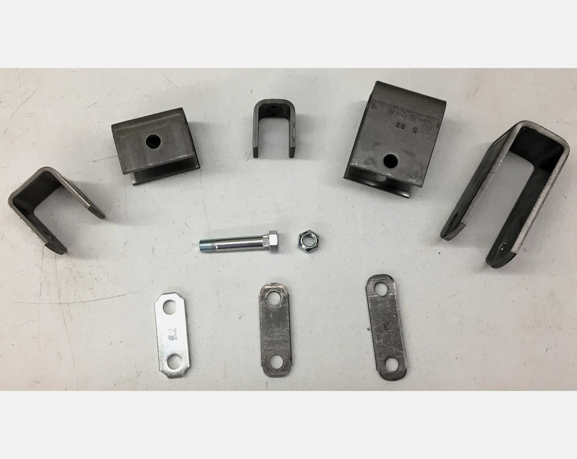 Metal trailer suspension hardware laid out on a white surface, including brackets, shackles, bolts, and a U-bolt.