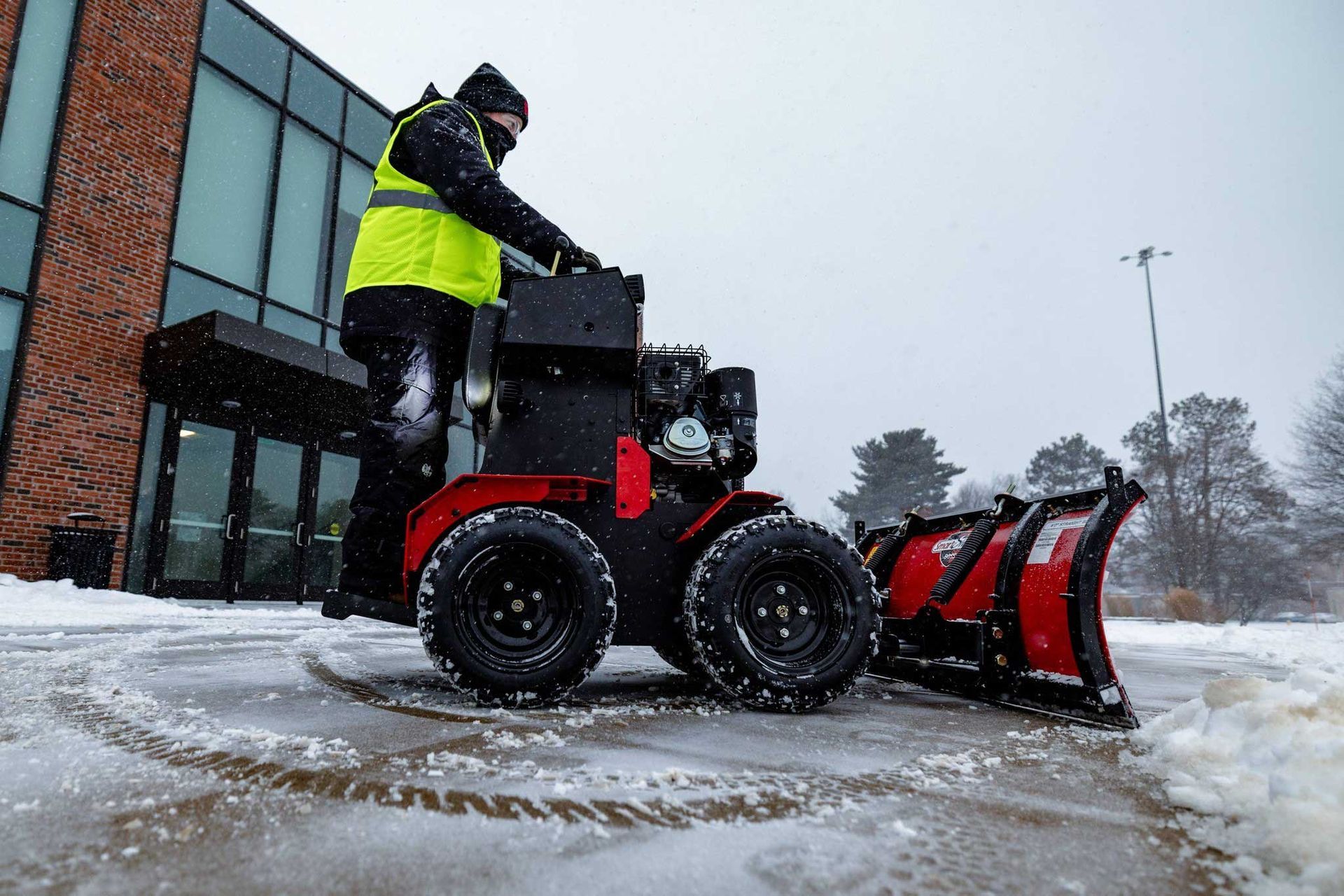 Person operating a small snow plow in front of a brick building during a snowfall. The person wears a yellow safety vest.