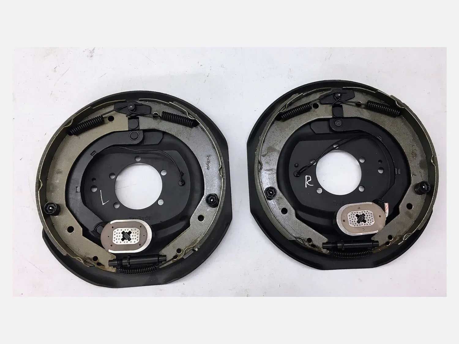 Two black trailer brake assemblies on a white surface, showing brake shoes, springs, and mounting hardware.
