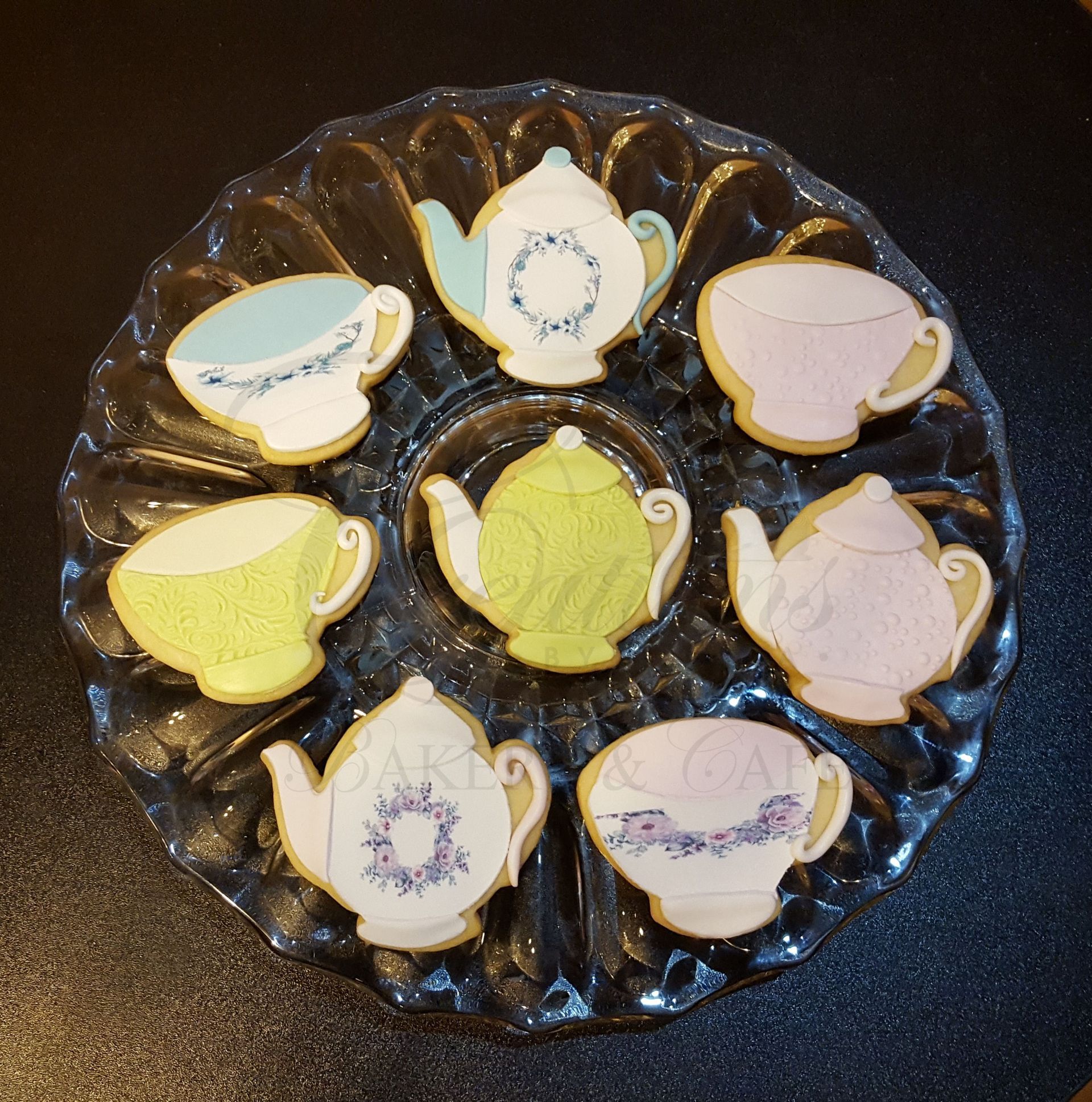 Assorted pastel teacup-shaped cookies arranged on a dark glass plate.