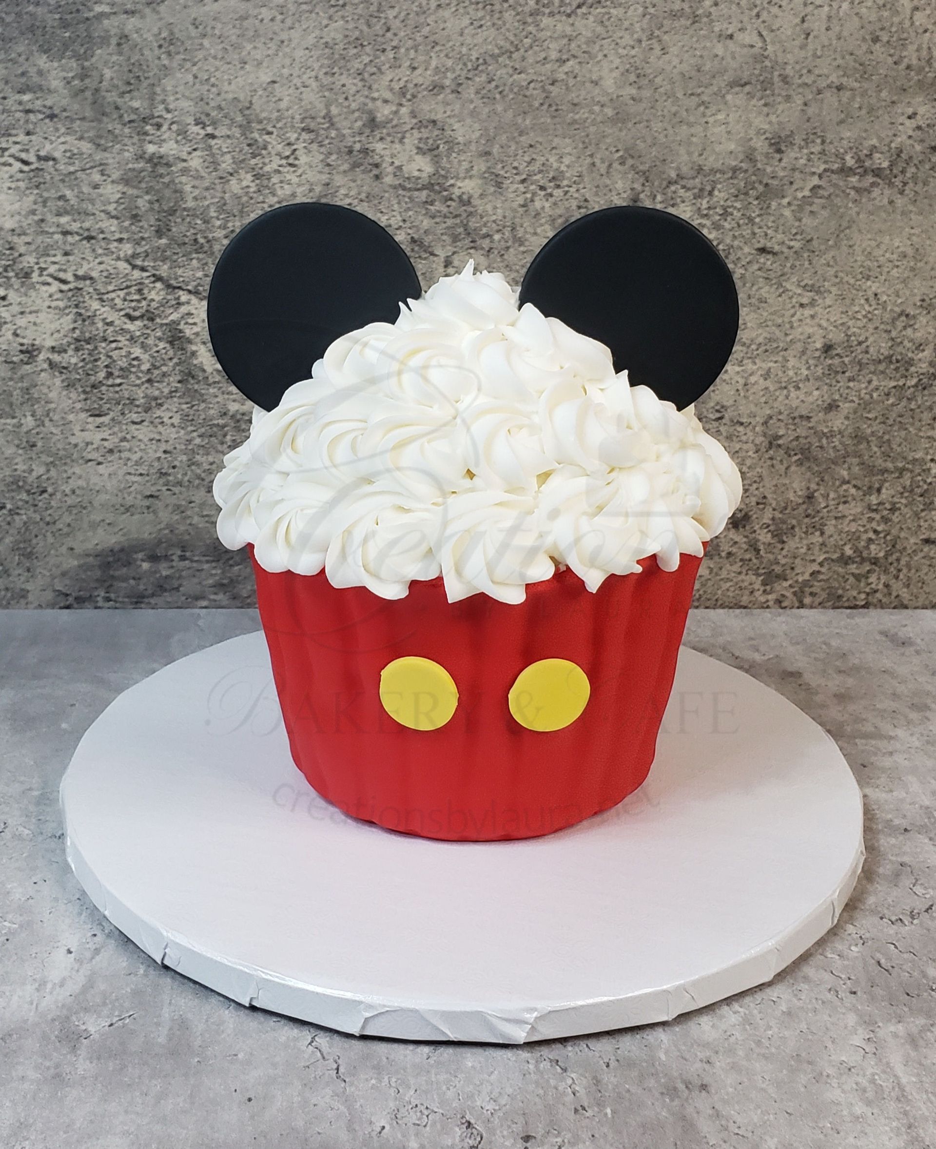 Red cupcake with white frosting and black mouse ears on a white stand, styled like Mickey Mouse.