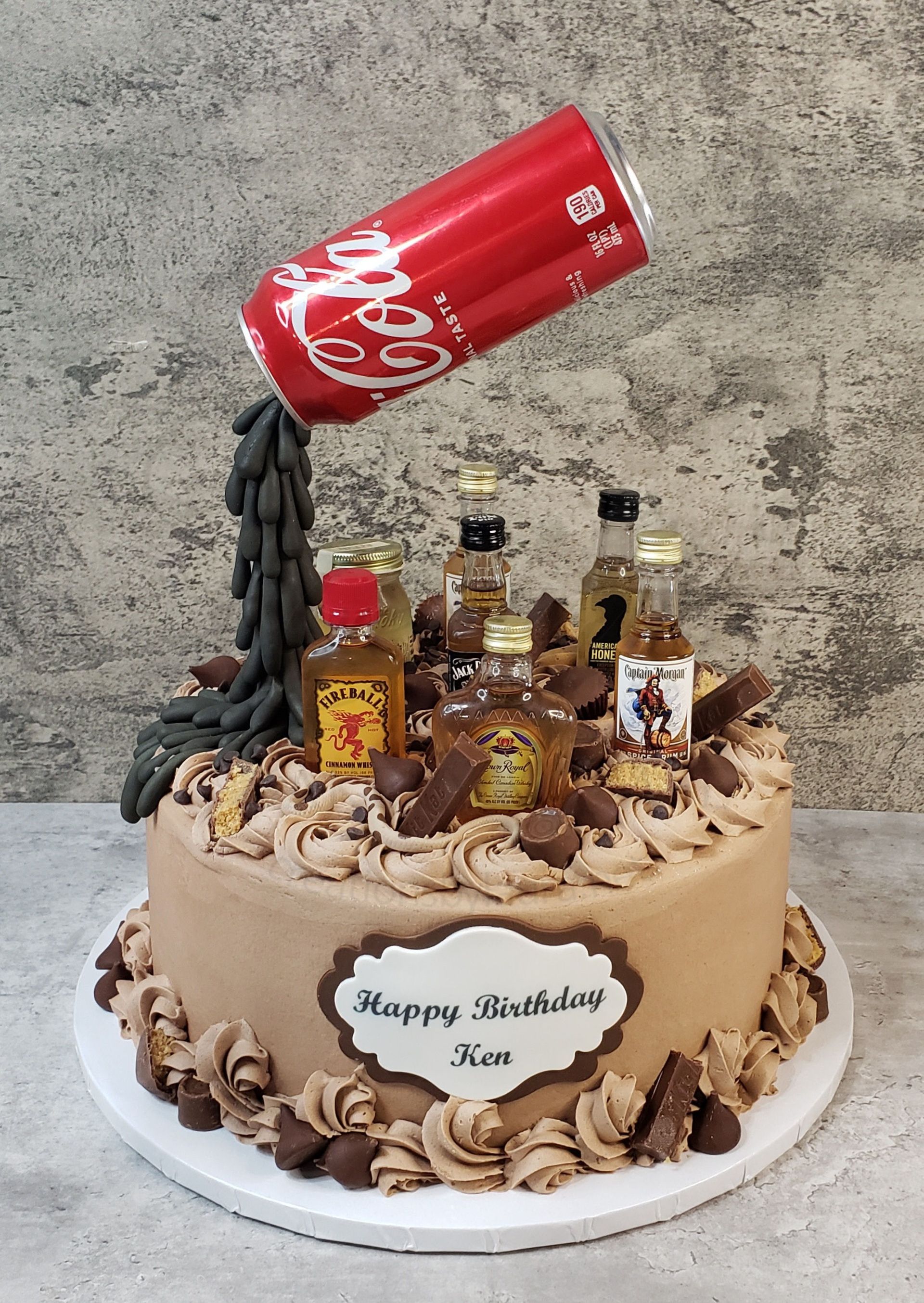 Birthday cake topped with a Coca-Cola can and liquor bottles, with “Happy Birthday” plaque