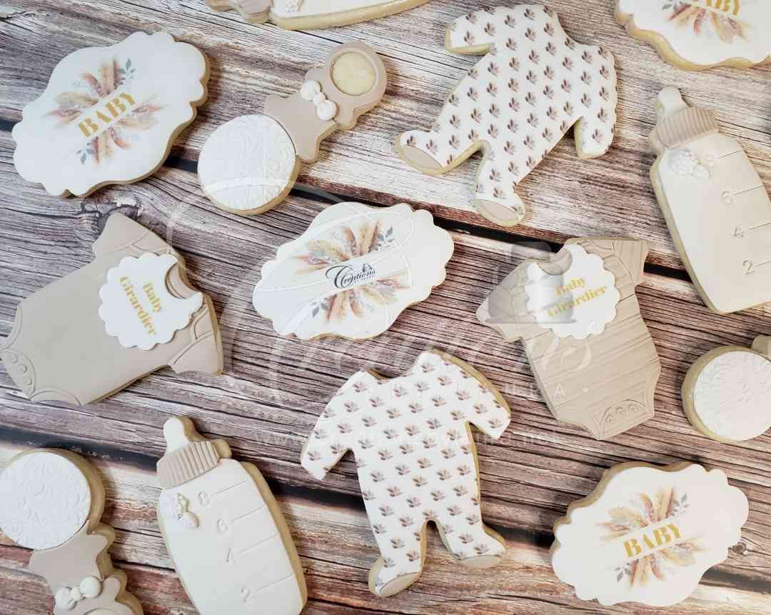 Boho-themed baby shower sugar cookies shaped like onesies, bottles, rattles, and plaques on a rustic wood background.