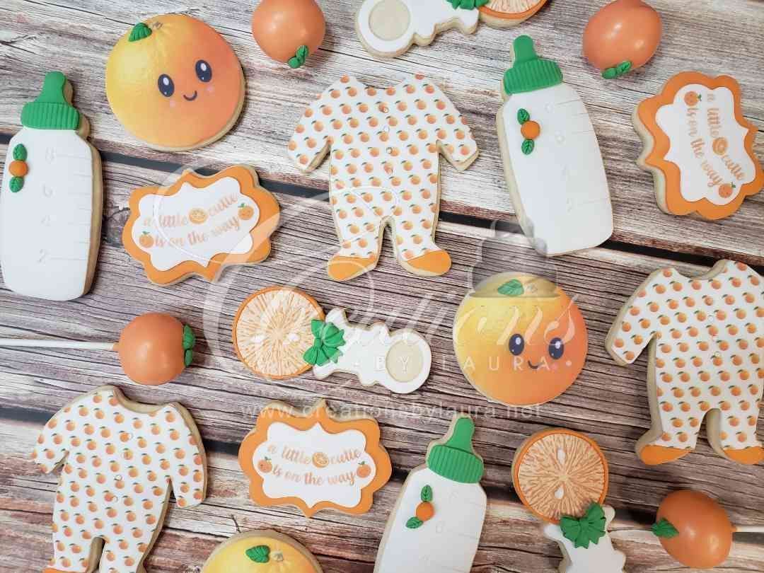 Baby-themed cookies, including bottles, patterned onesies, round orange faces, and plaques on a rustic wooden background.
