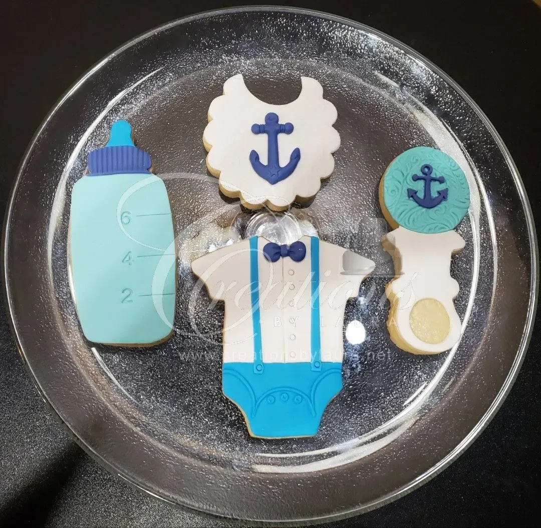 Four baby-themed sugar cookies—a bottle, bib, onesie, and rattle—arranged on a clear plate with a nautical anchor motif.