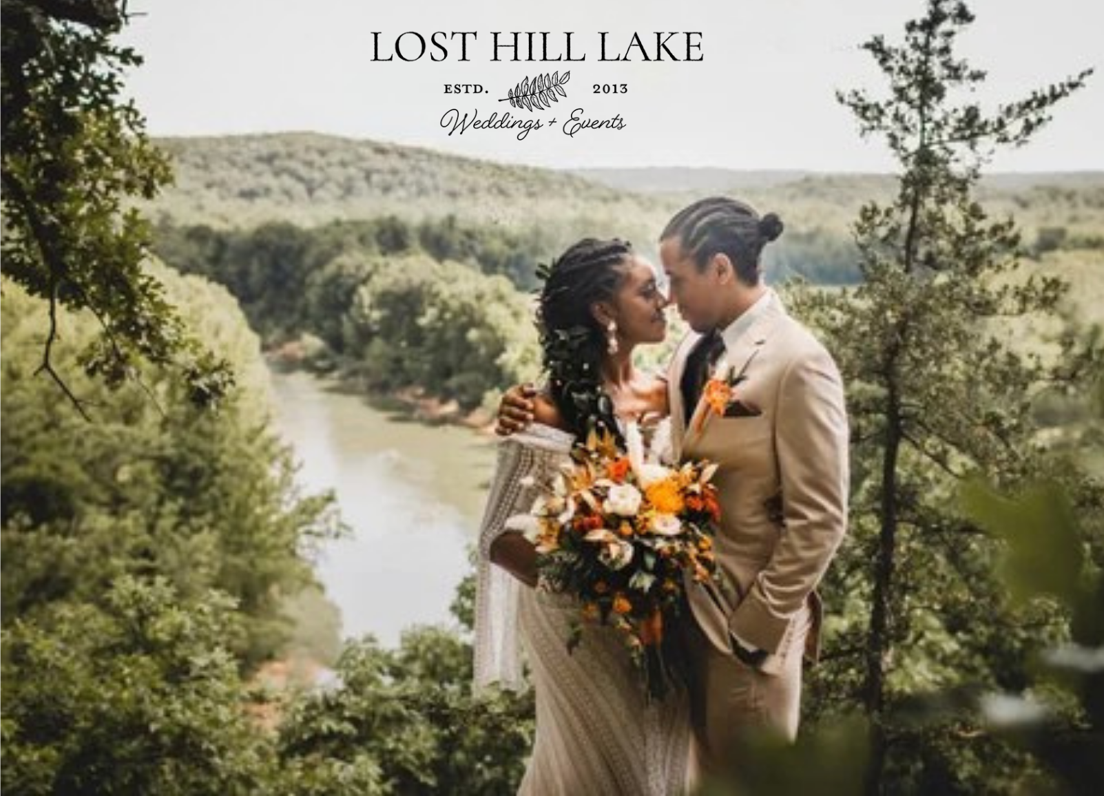 Couple in wedding attire standing on a hill overlooking a lake, holding a bouquet amid trees.