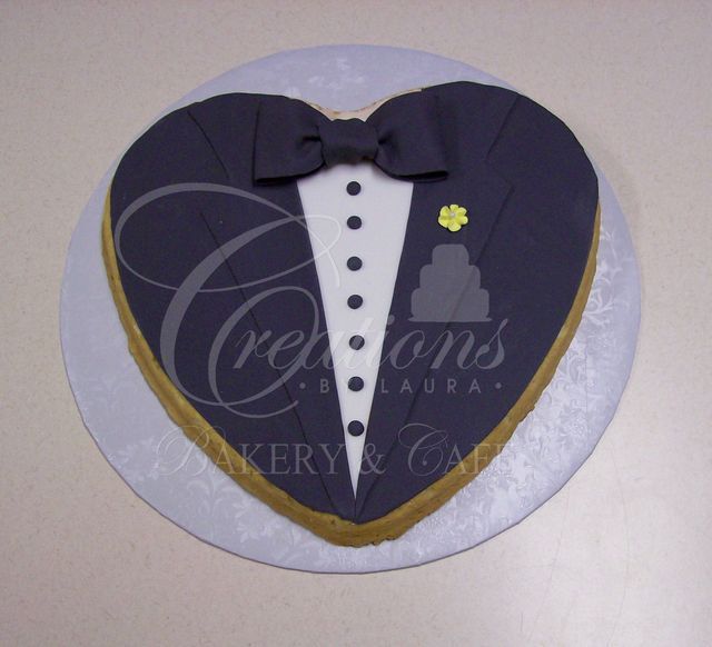 A heart-shaped cake decorated to look like a dark suit jacket with a white shirt, black bowtie, and small yellow flower.