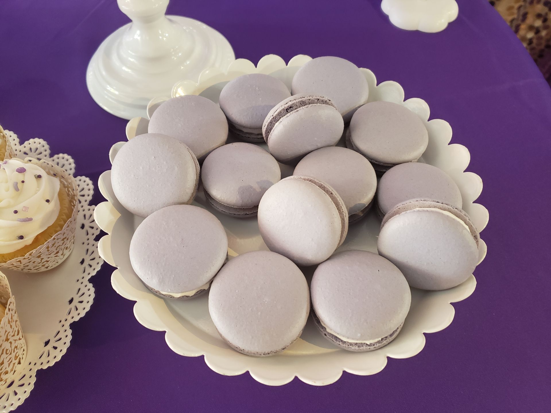 A white scalloped plate filled with light purple macarons sitting on a purple tablecloth next to a frosted cupcake.