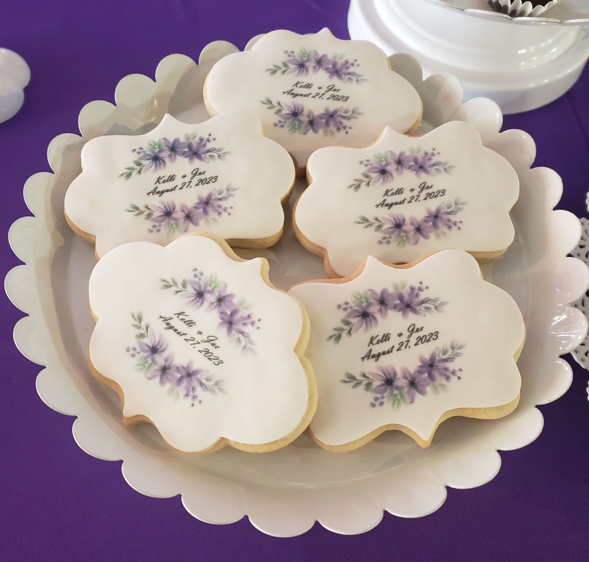 Five scalloped-edge sugar cookies on a white platter, each printed with purple floral wreaths and the text