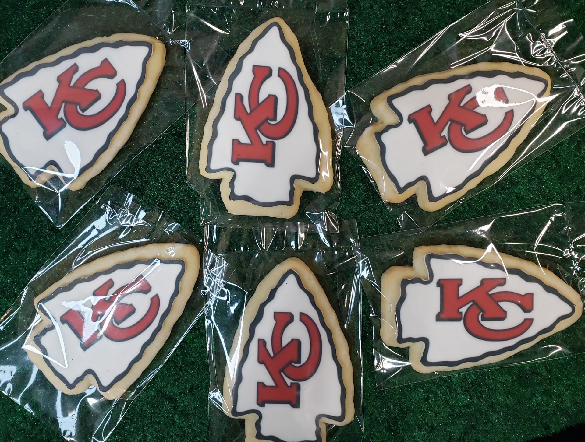 Six Kansas City Chiefs arrowhead-shaped cookies in clear plastic wrappers arranged on a green surface.