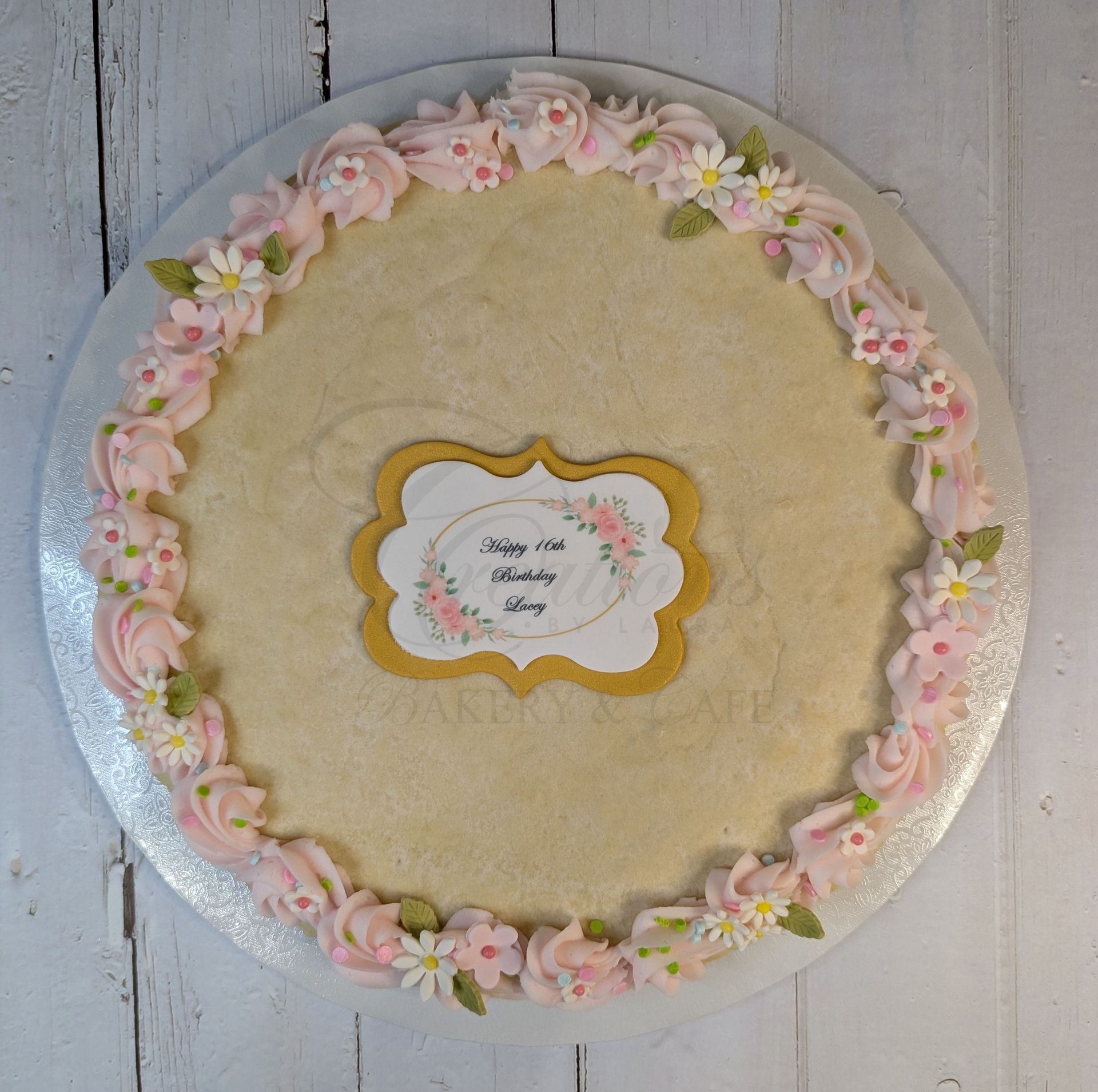 Round beige decorative cake with pink and white floral frosting border and a small center label on a gray wood background