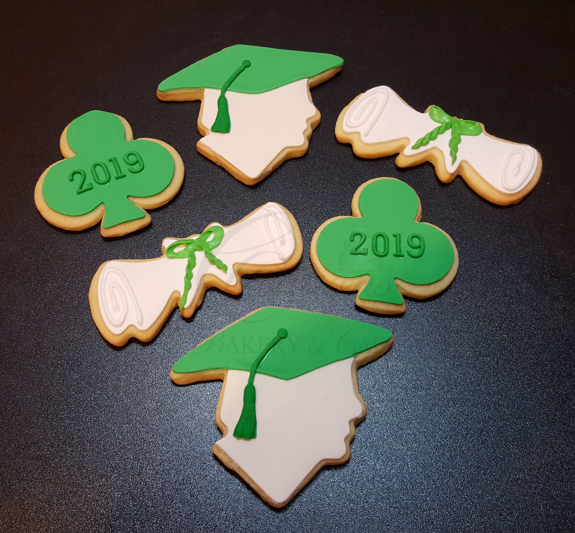 Graduation-themed decorated cookies with green caps, diplomas, and “2019” clovers on a dark surface