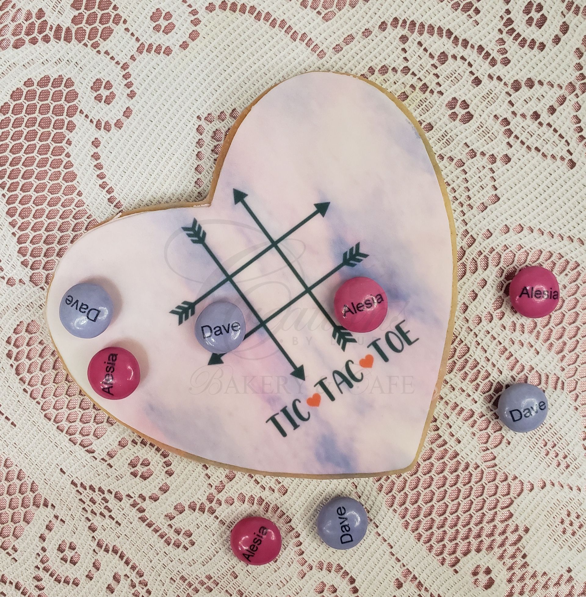 Heart-shaped tic-tac-toe board on lace, with pink, blue, and black game pieces scattered around.