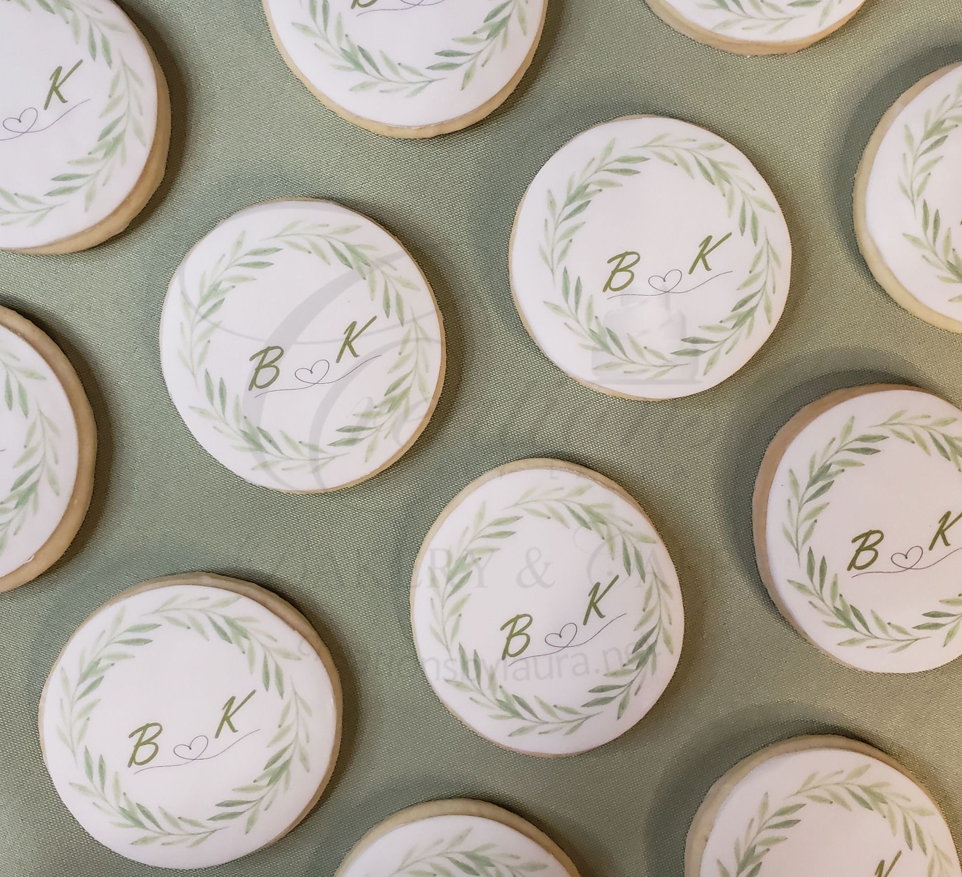 Round white cookies with green wreath designs and gold initials, arranged on a textured green cloth