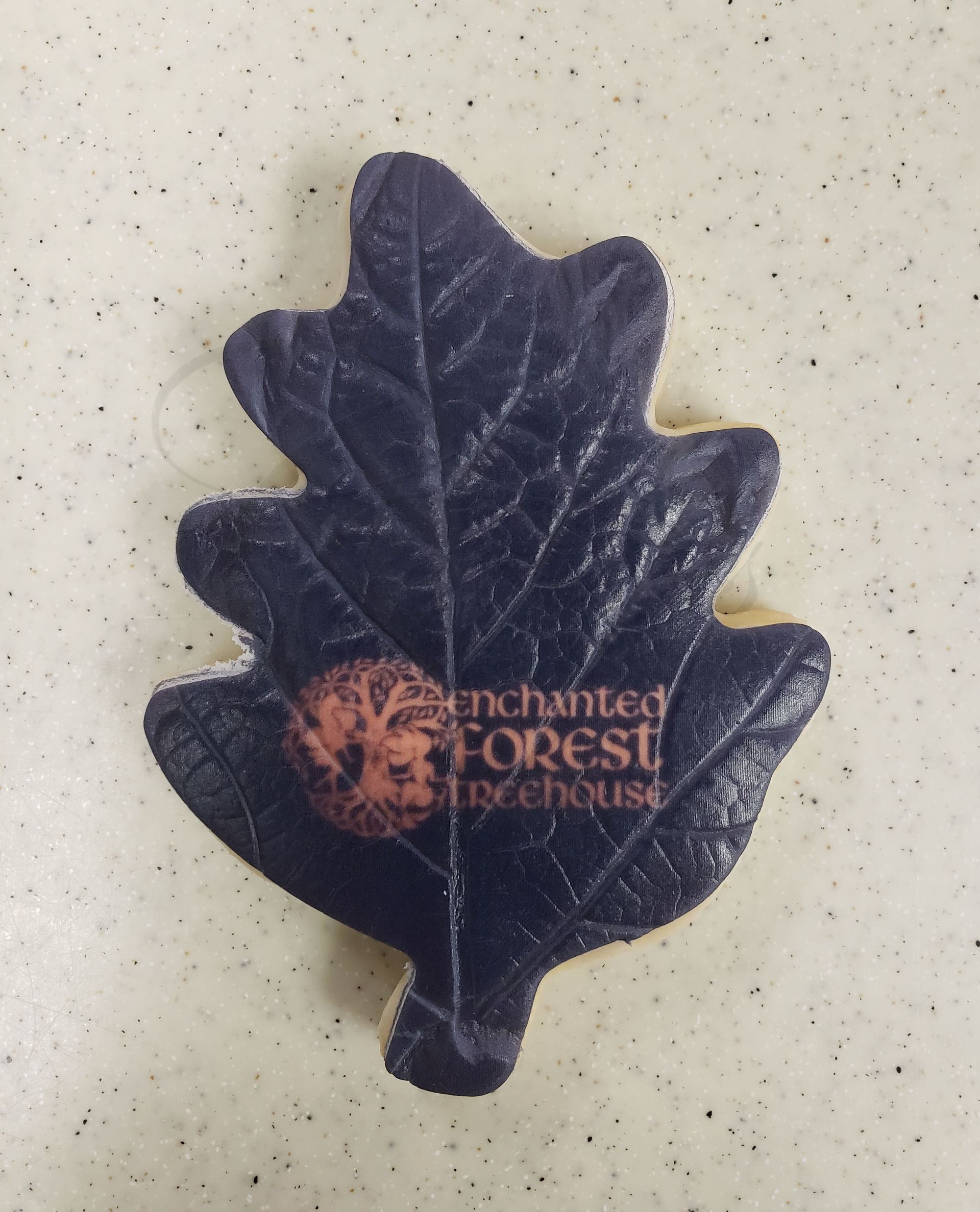 Dark purple leaf-shaped embroidered patch with a small orange cat face and faint text on a beige surface.