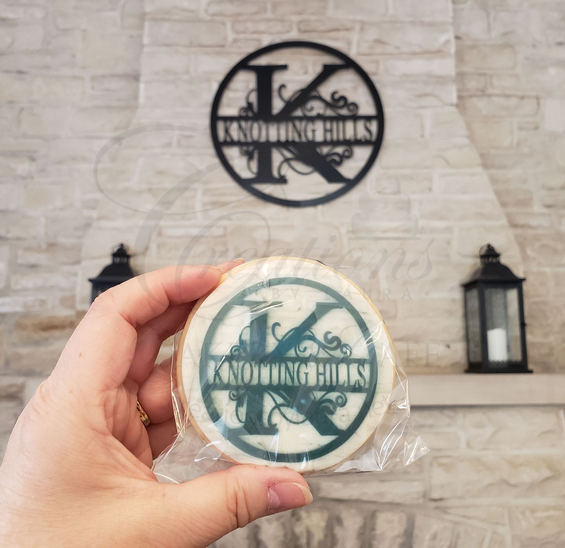 Hand holding a wrapped round cookie with a black Knoxville logo in front of a matching wall sign.