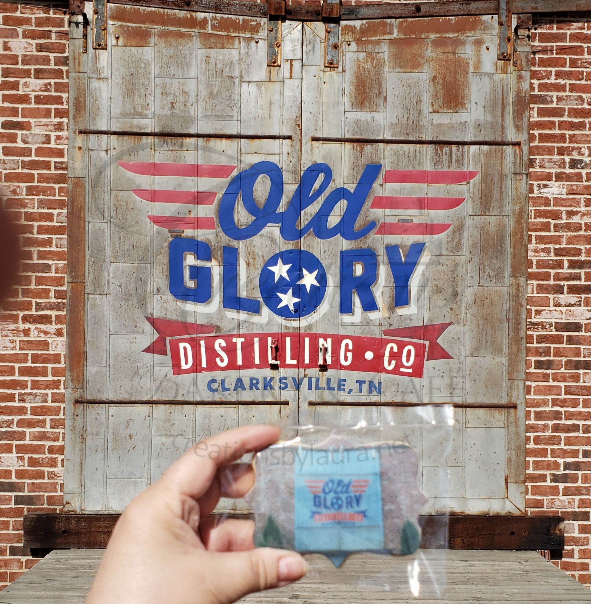 Hand holding a small bottle in front of an Old Glory Distilling Co. mural on a brick wall.