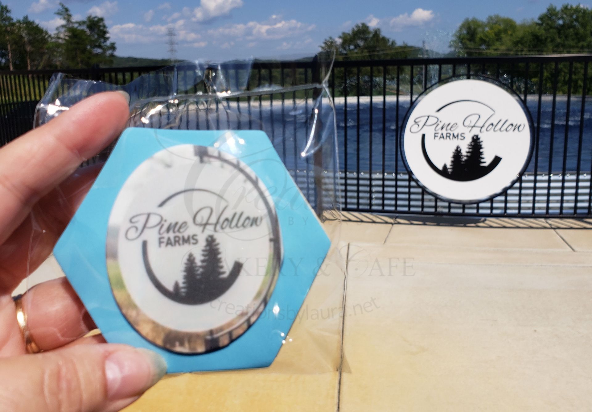 Hand holding a blue hexagonal keychain in front of a gated farm entrance with a round logo sign.