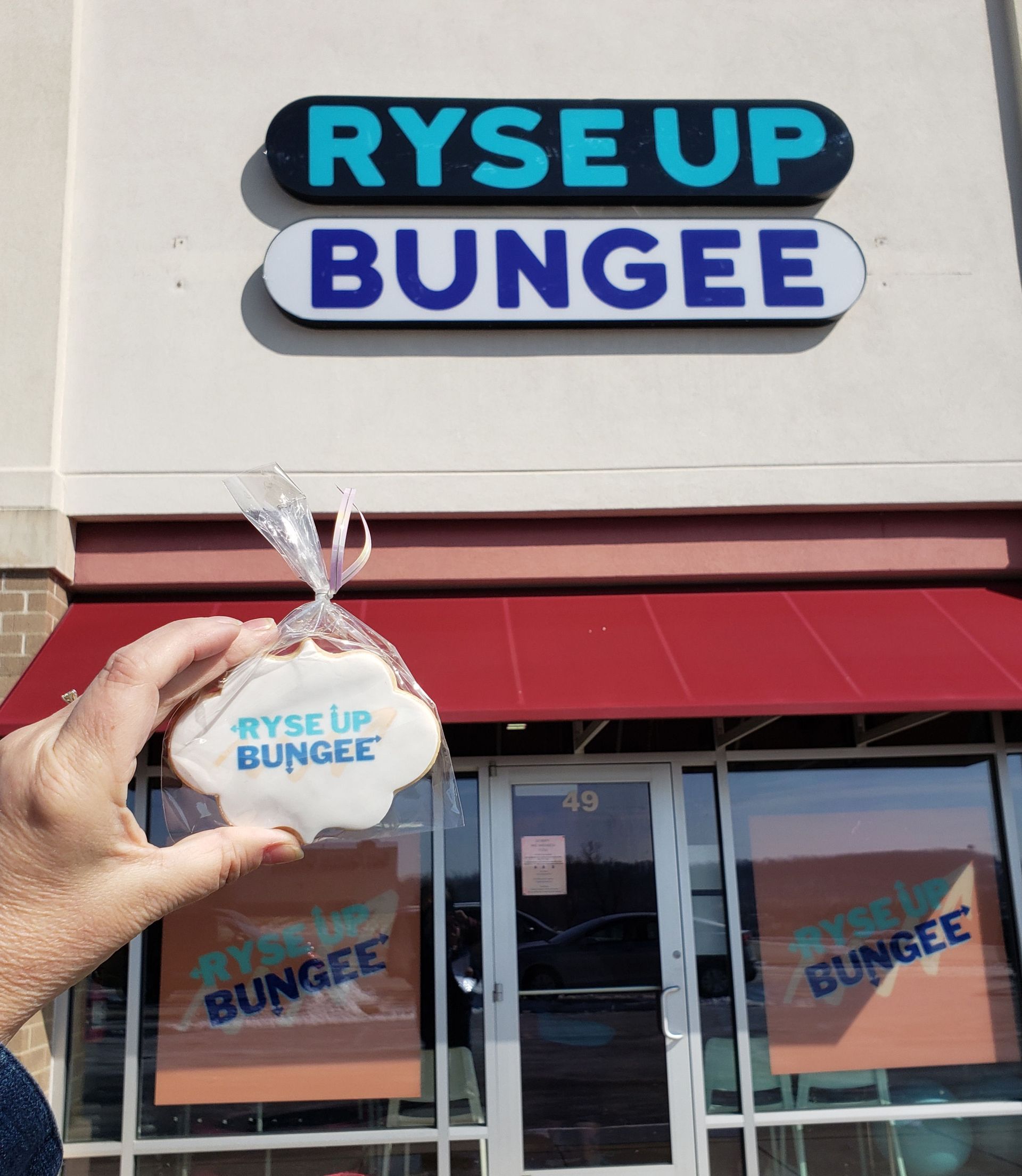 Hand holding a clear cup in front of the Ryse Up Bungee storefront sign.