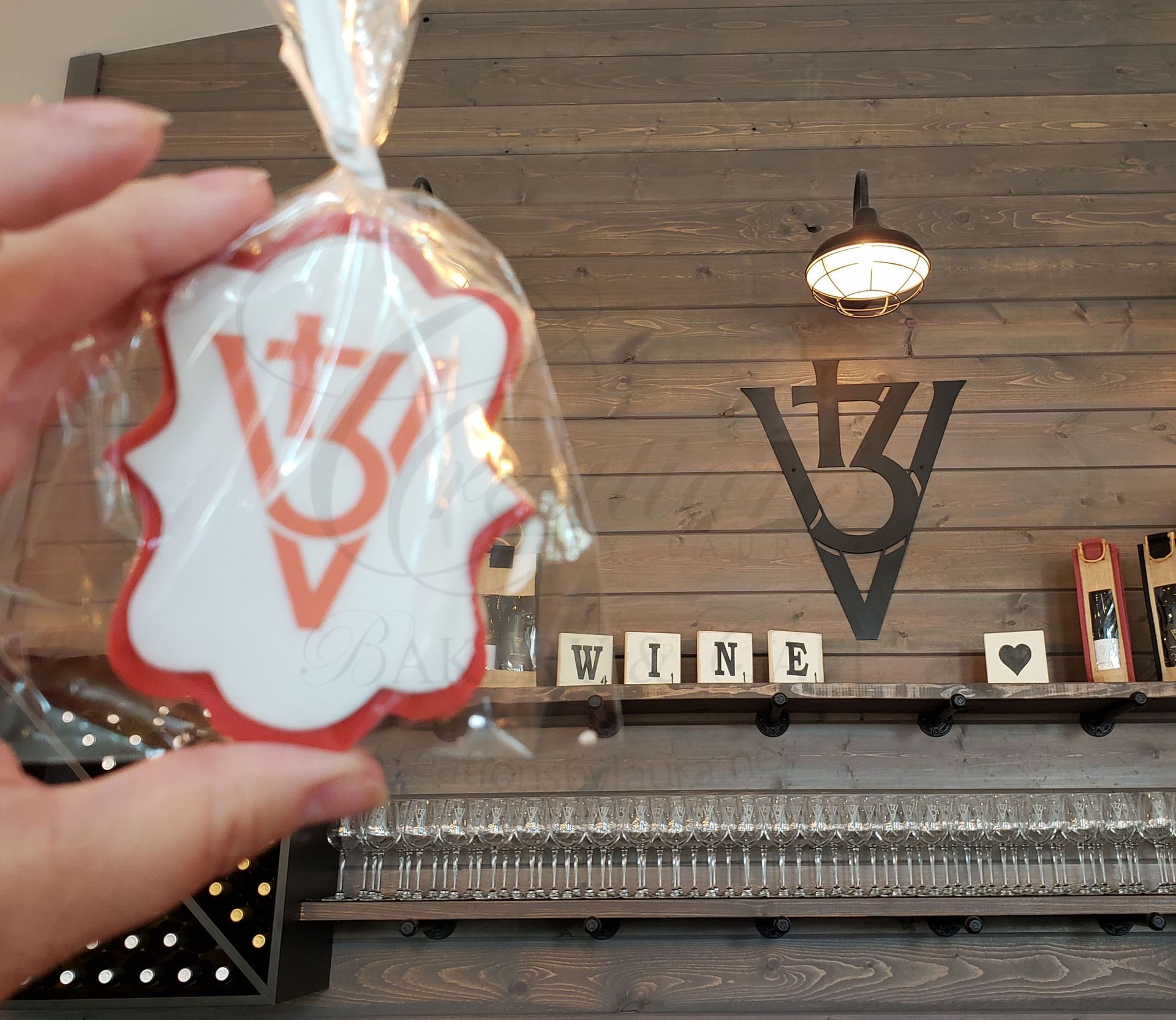 Hand holding a small wrapped tag in front of a rustic wine shop wall with a large wine logo and shelf decor.