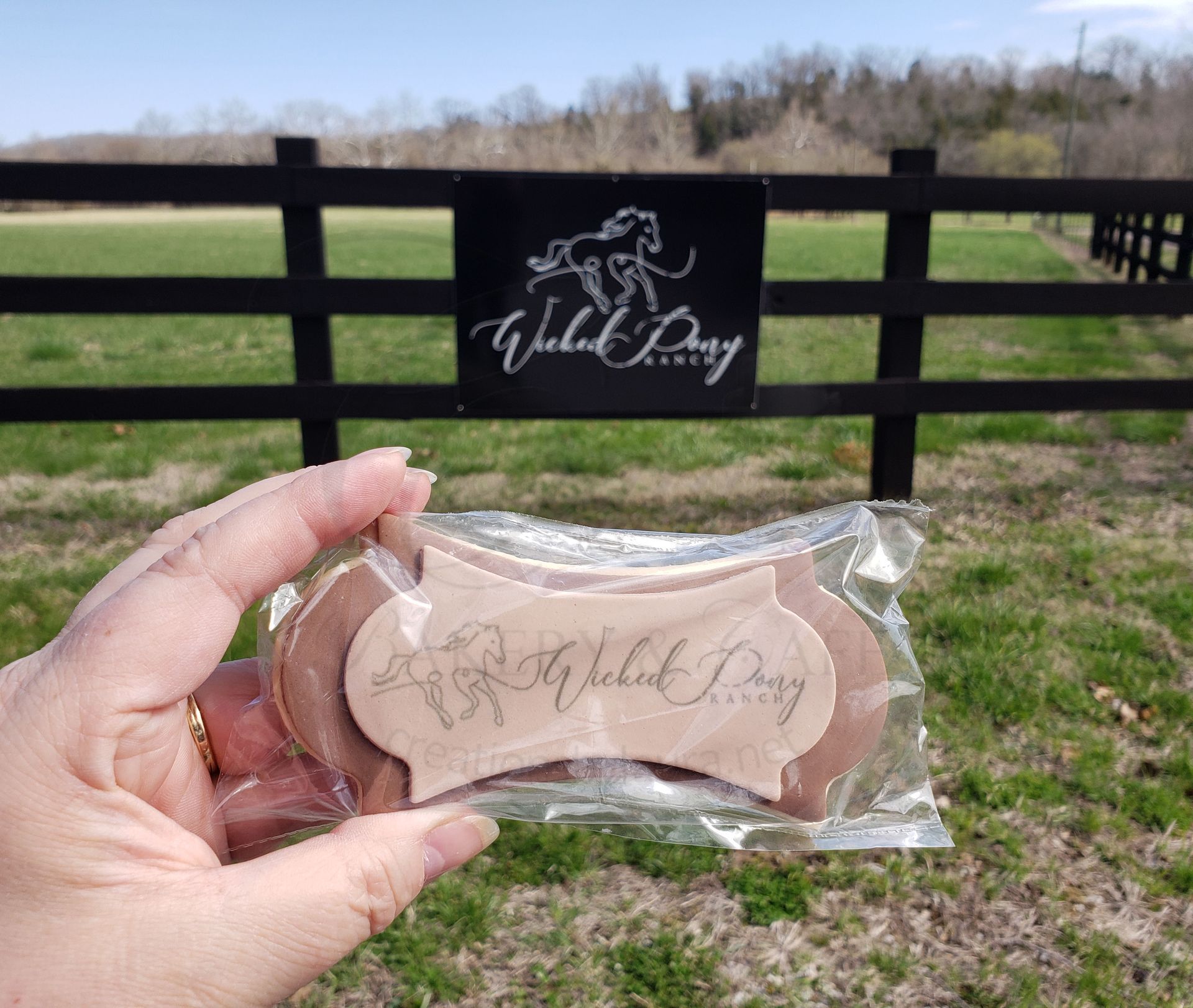 Hand holding a clear-wrapped pink soap bar in front of a fenced grassy field and a branded sign.