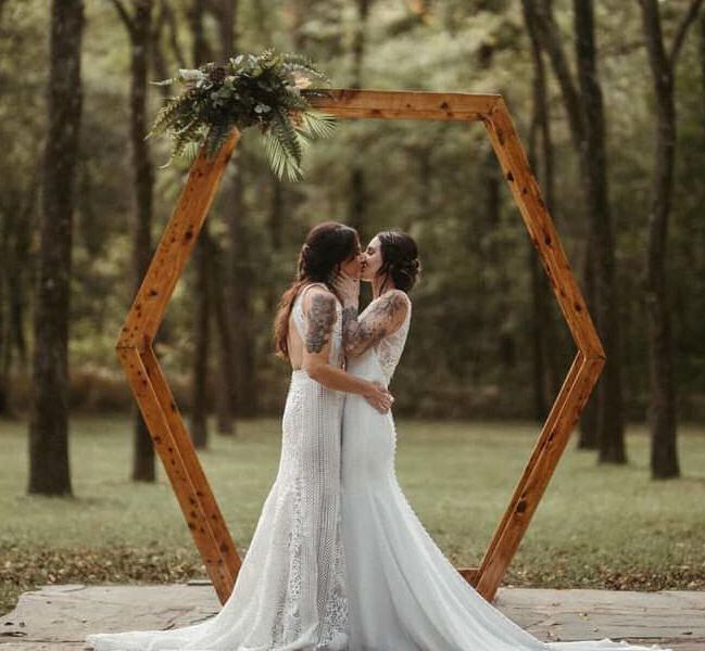Two brides are kissing under a wooden arch in the woods