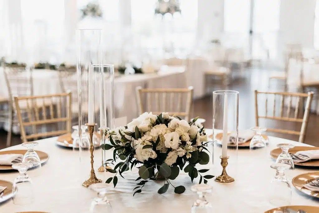 Round table set for a formal event, featuring floral centerpiece, gold accents, and transparent candle holders.