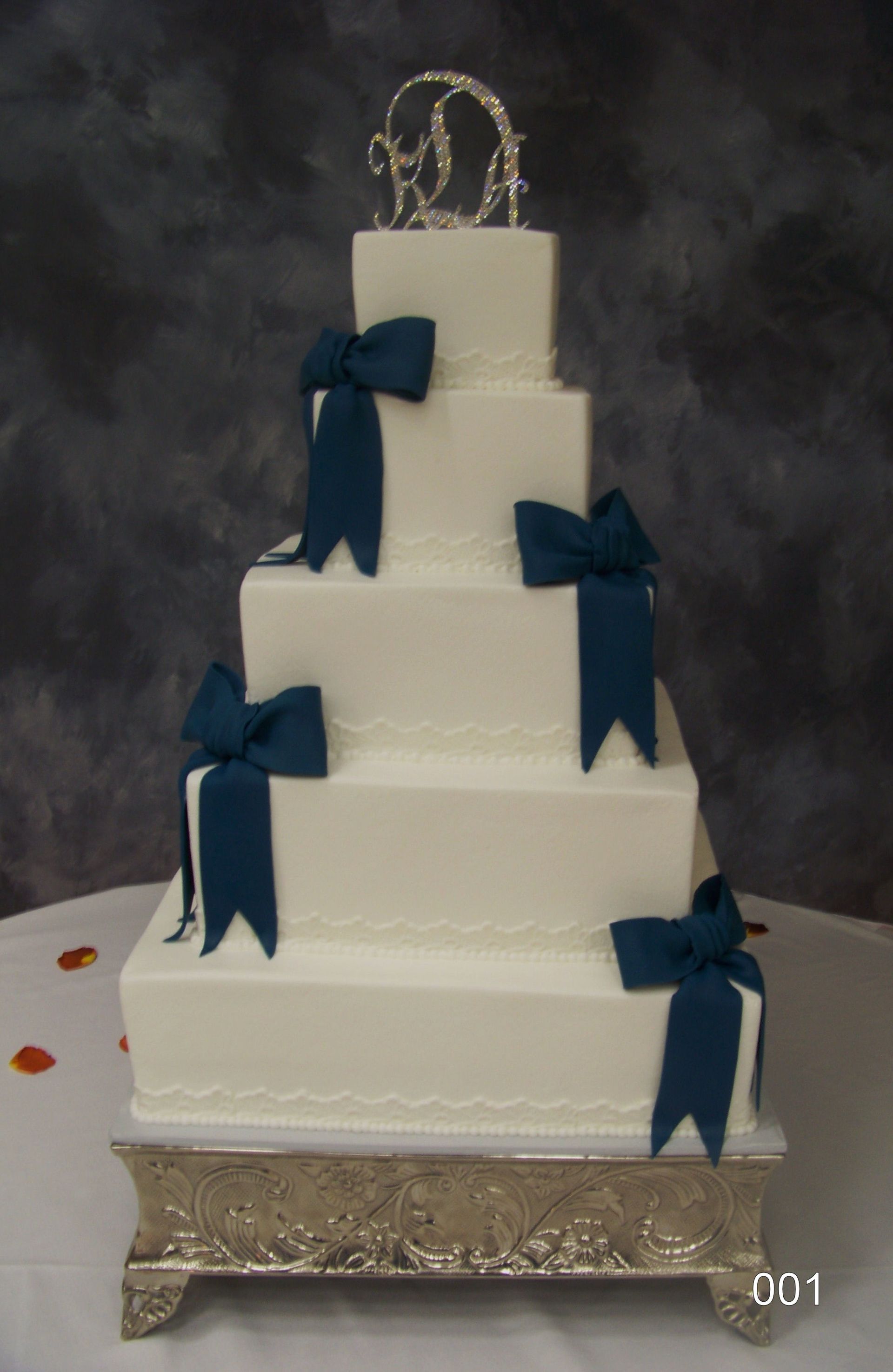 Four-tier white wedding cake with navy blue ribbon bows on a silver tablecloth and decorative topper